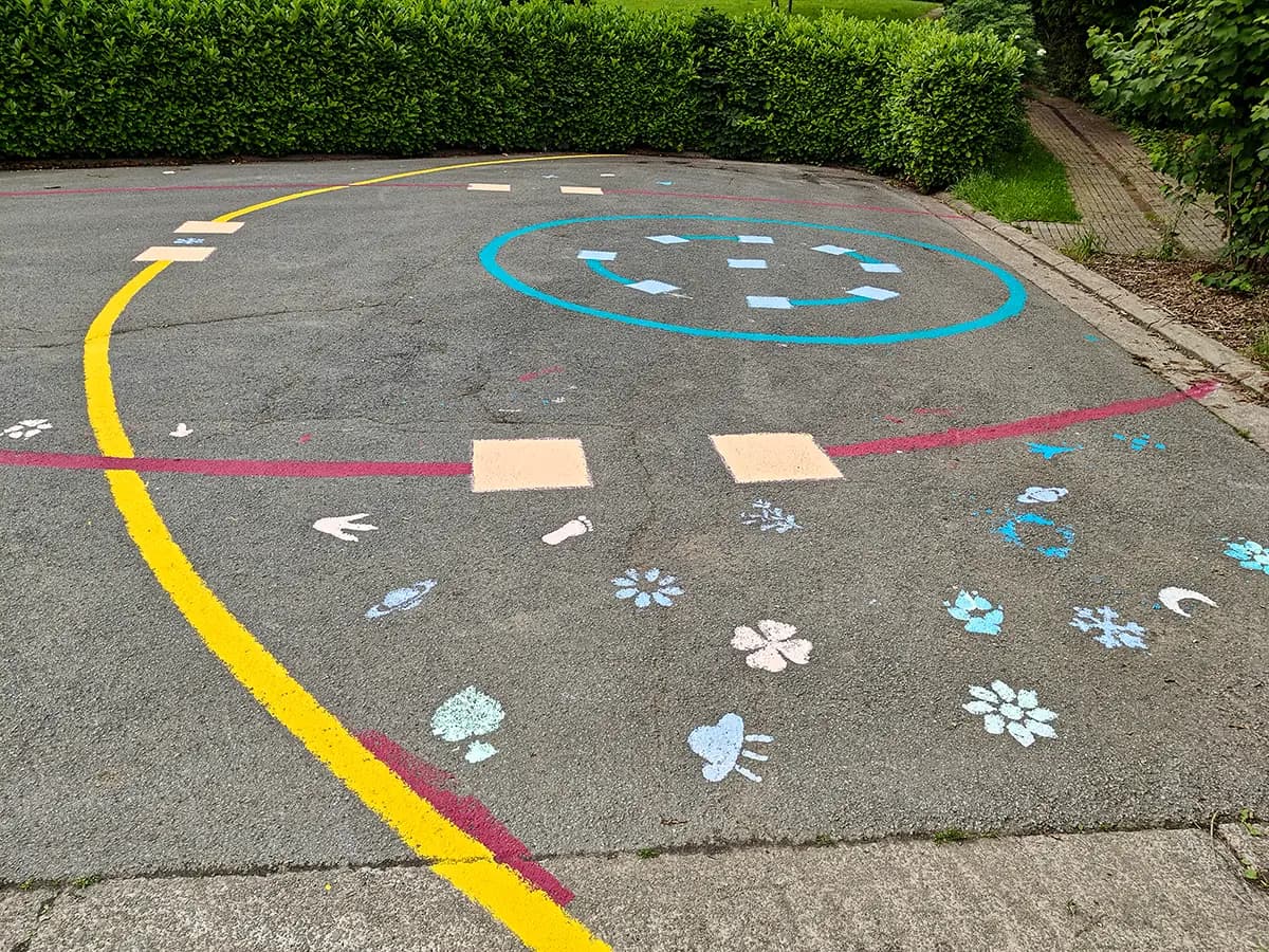 Speelplein op asfalt met een gele boog, een rode middellijn met twee vakken, een blauwe cirkel en gestencilde bloemen.