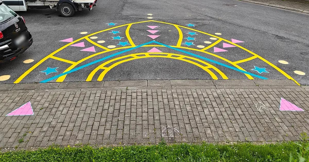 Gele boog op het asfalt met roze driehoeken, blauwe sterren en gele stippen. In het midden komt later een basketbalring.