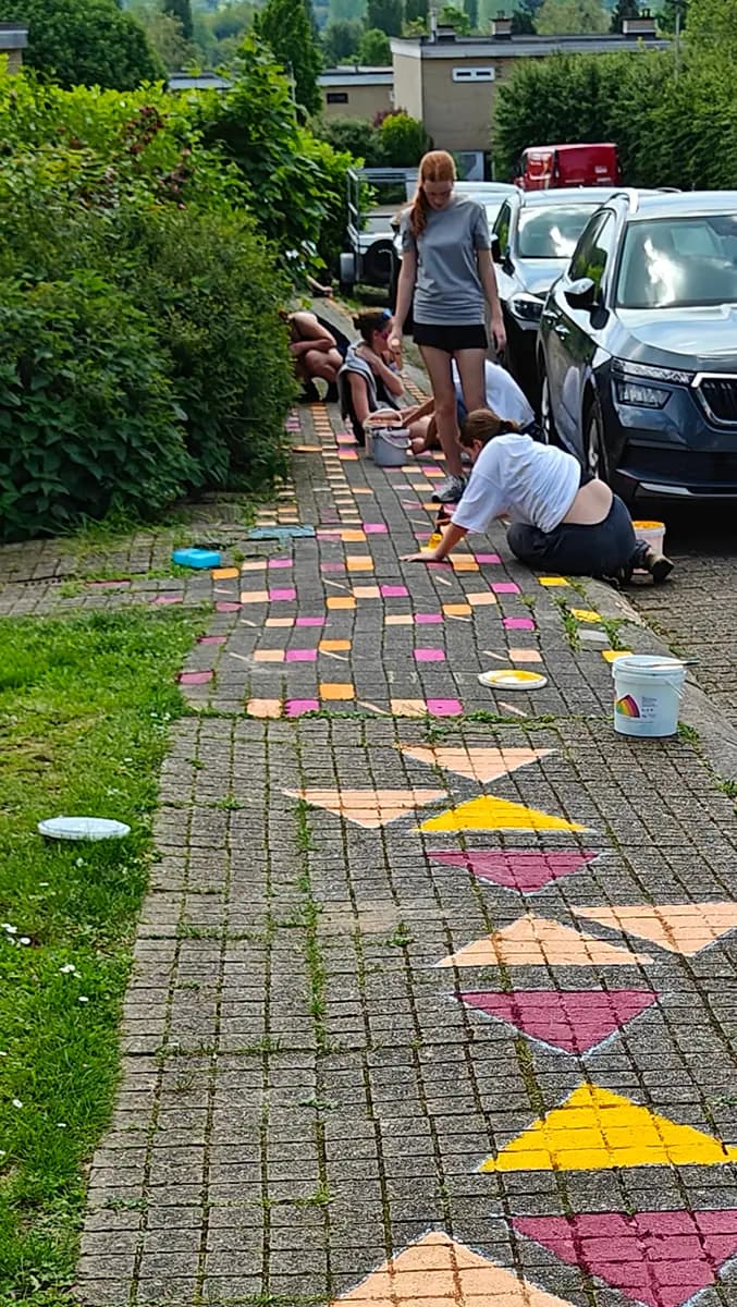 Jongeren van de jeugdbeweging schilderen gekleurde vakjes op het voetpad. Vooraan zijn de driehoeken al klaar.